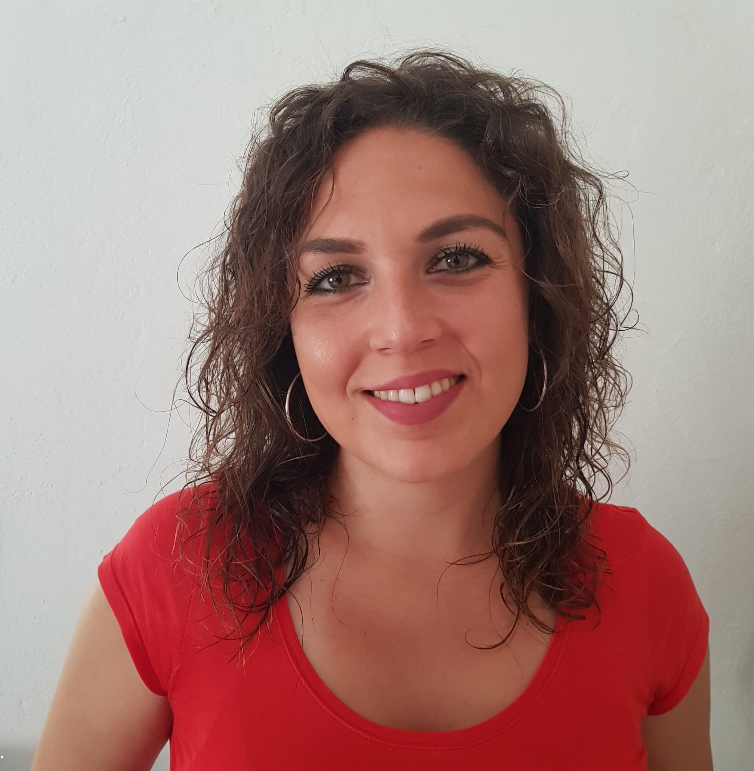 Mujer con cabello rizado y aretes grandes, sonriendo, lleva una camiseta roja. Fondo blanco liso.
