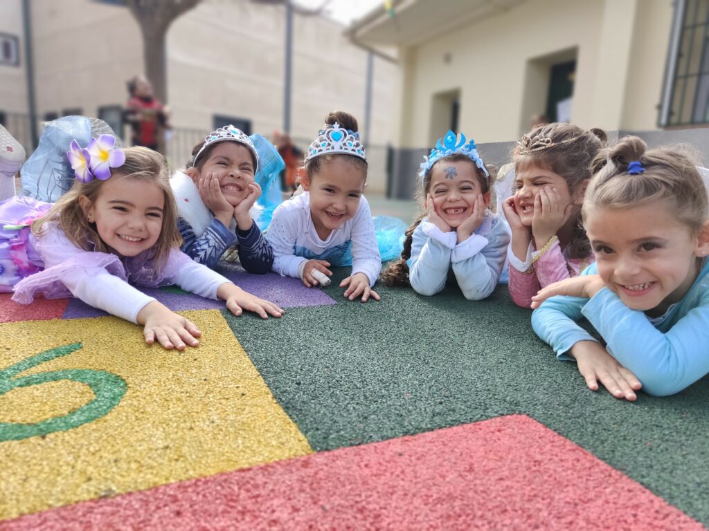 Niñas disfrazadas con coronas sonríen acostadas en el suelo de un patio de juegos al aire libre.