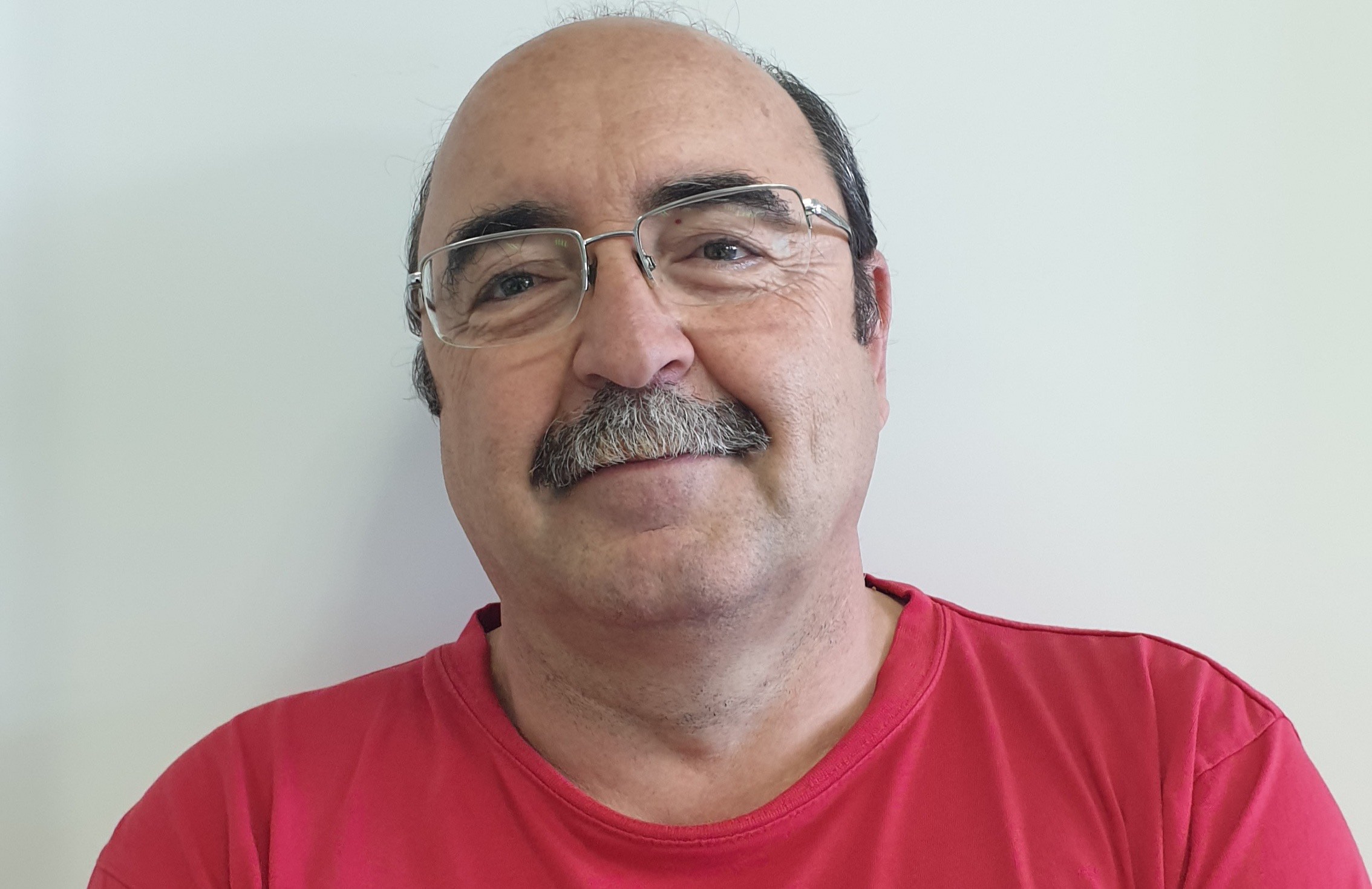 Hombre de mediana edad con gafas y bigote, vistiendo camiseta roja, sonriendo ligeramente frente a un fondo blanco.