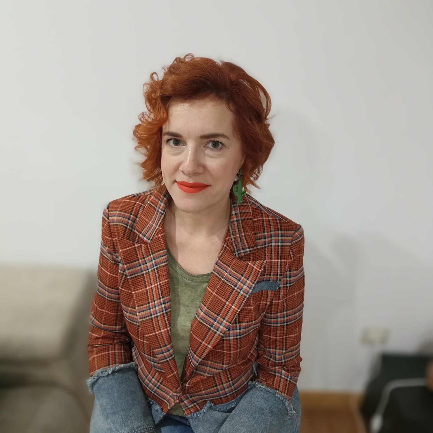 Mujer con cabello rojo rizado, lleva chaqueta de cuadros rojos y verdes, camiseta verde y pendientes verdes.