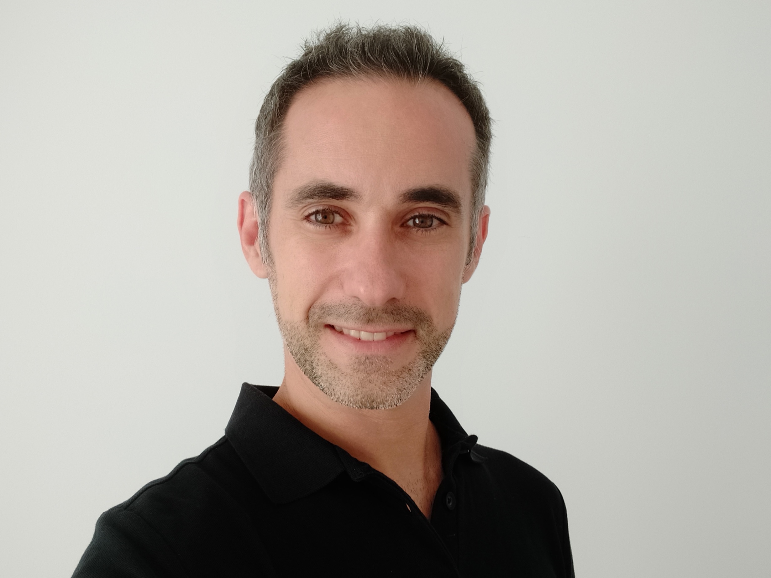 Hombre con barba corta y cabello canoso, sonriendo, vestido con camisa negra, fondo blanco.