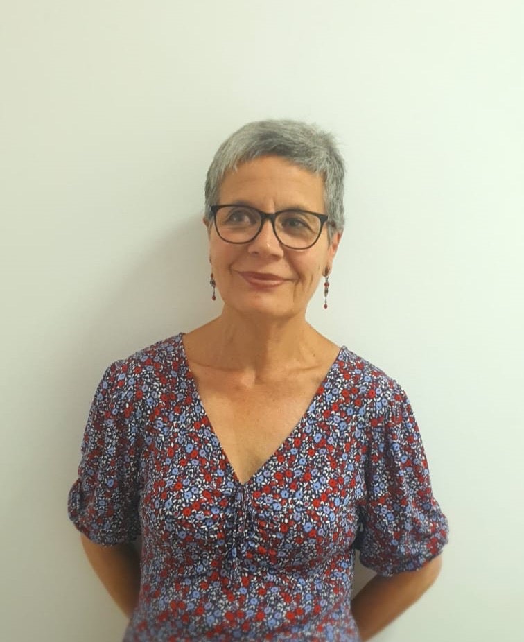 Mujer de cabello corto gris con gafas y vestido floral sonríe frente a un fondo blanco.