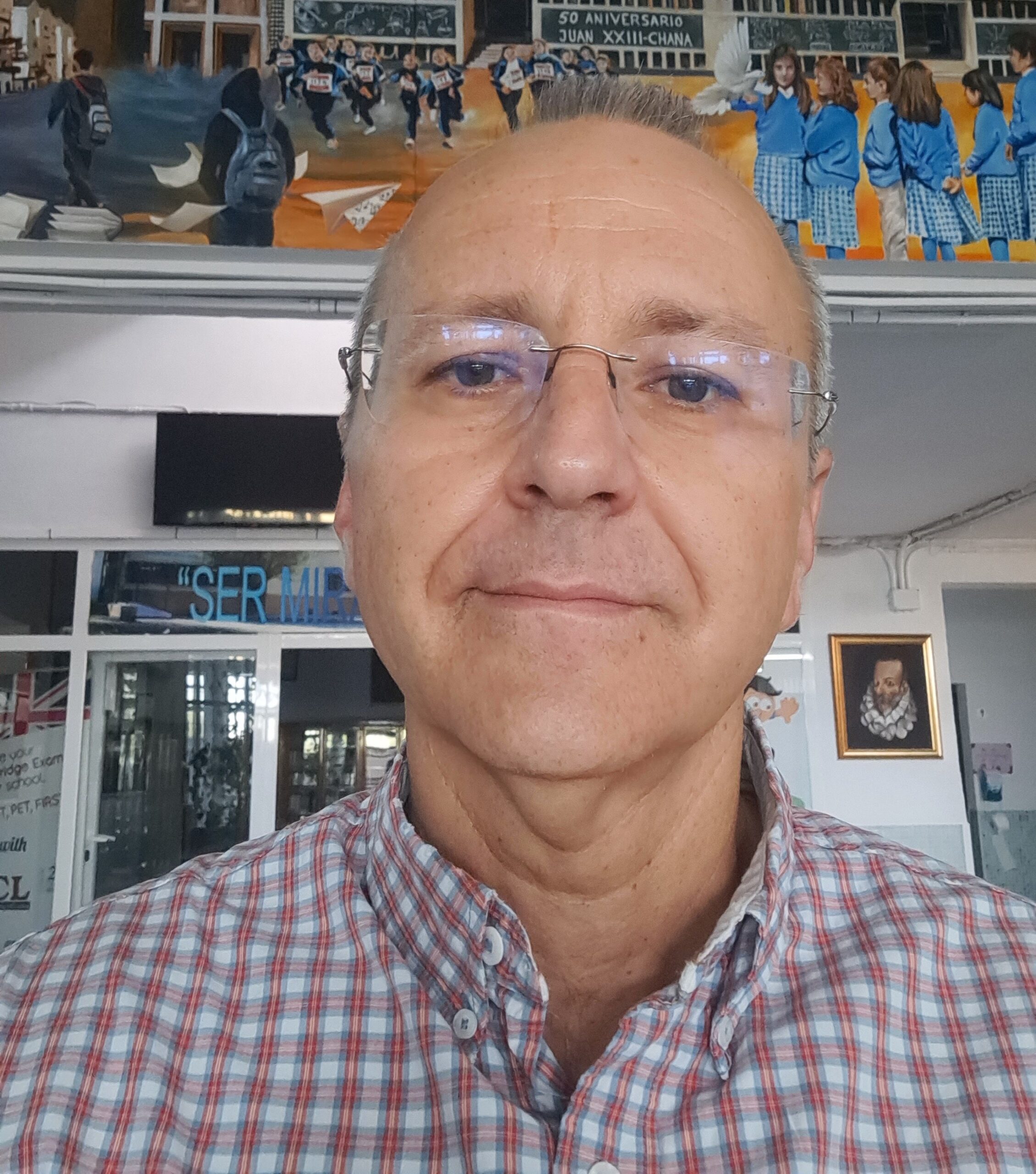 Hombre de mediana edad con camisa a cuadros, de pie en un espacio interior con fondo de mural y retrato enmarcado.