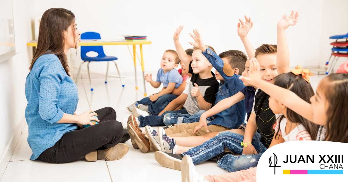 Maestra sentada frente a niños en el aula, varios levantan la mano. Logo de Juan XXIII Chana en la esquina.